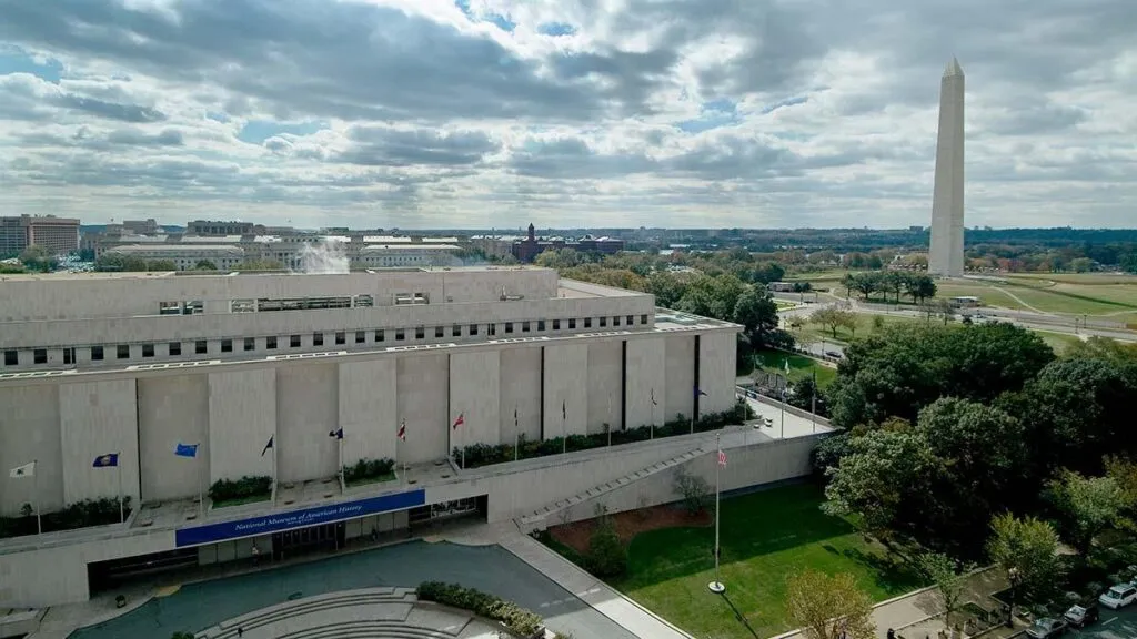 A long pale stone federal building seen from the air, a tall white obelisk on the skyline beyond it
