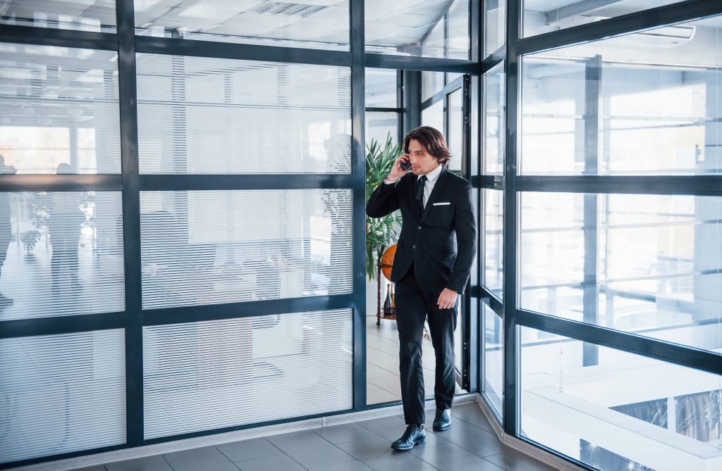 Man in suit walks by office glass walls, consulting Commercial Glass Repair needs on his phone | American Glass and Door